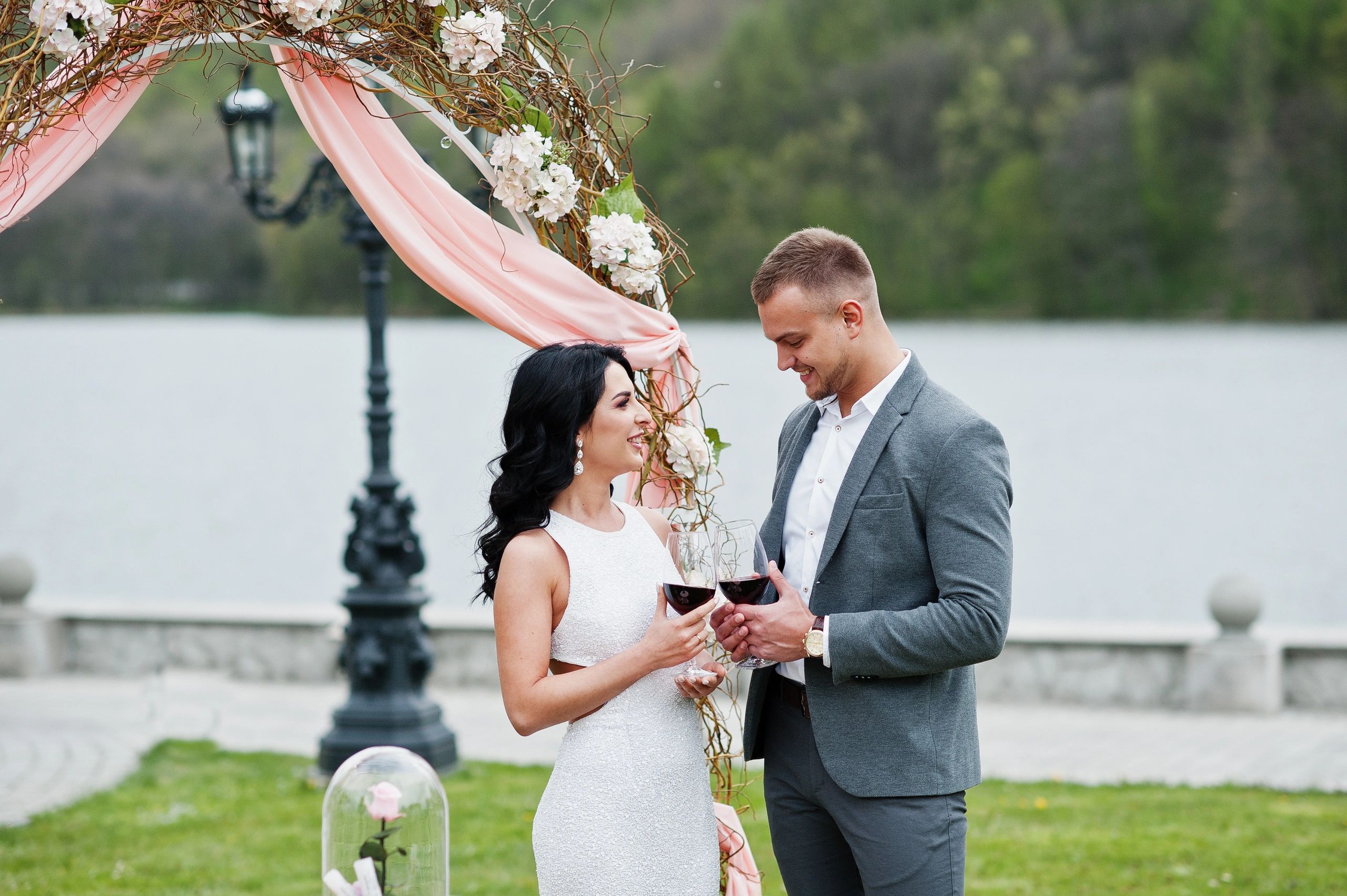 Young fashionable lovely couple against decor arch on wedding anniversary and marriage proposal. Glasses of vine at hands.